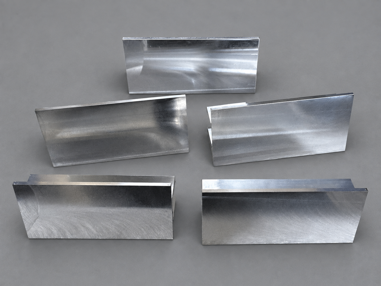 Five machined metal components showing flat faces, stepped edges, and visible tool marks